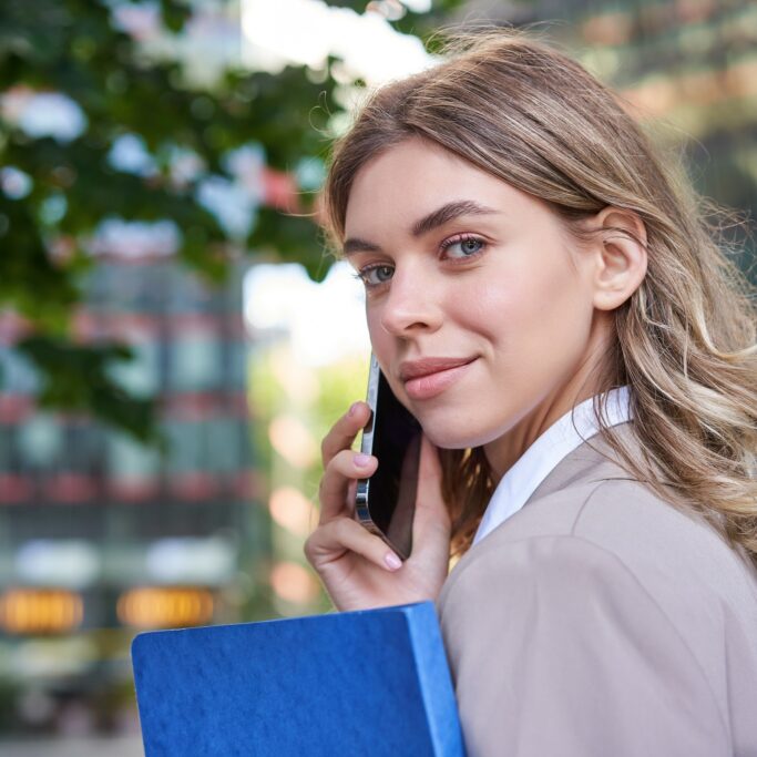 Close up portrait of corporate woman calling on mobile phone, walking on street with work documents in hand, wearing business suit